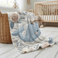 This embroidered look baby blanket features a gentle whale with a delicate bow, surrounded by soft floral elements in calming blue, cream, and natural tones.